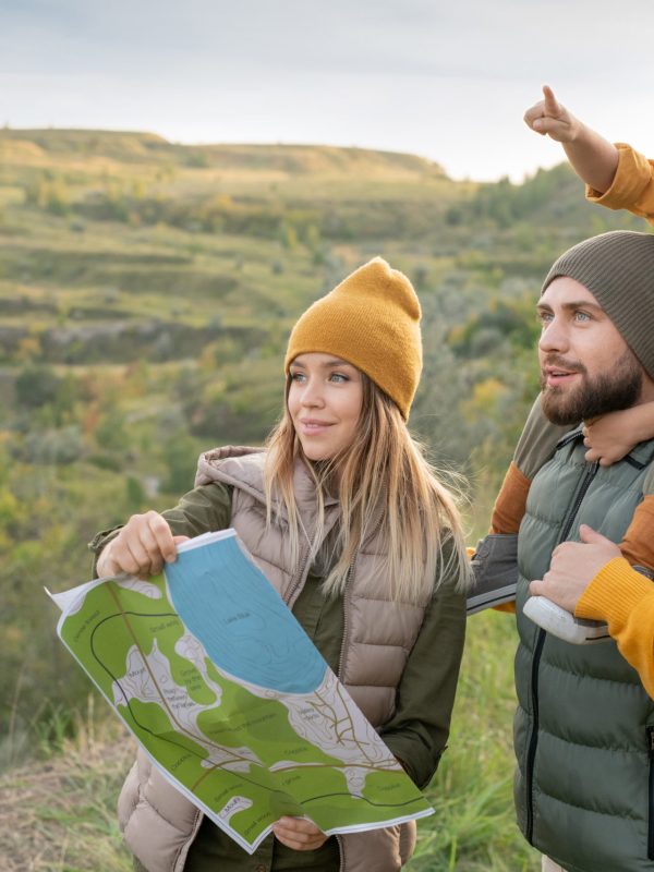 Happy young family of backpackers with map looking forwards while little boy pointing at far place against mountains during weekend trip